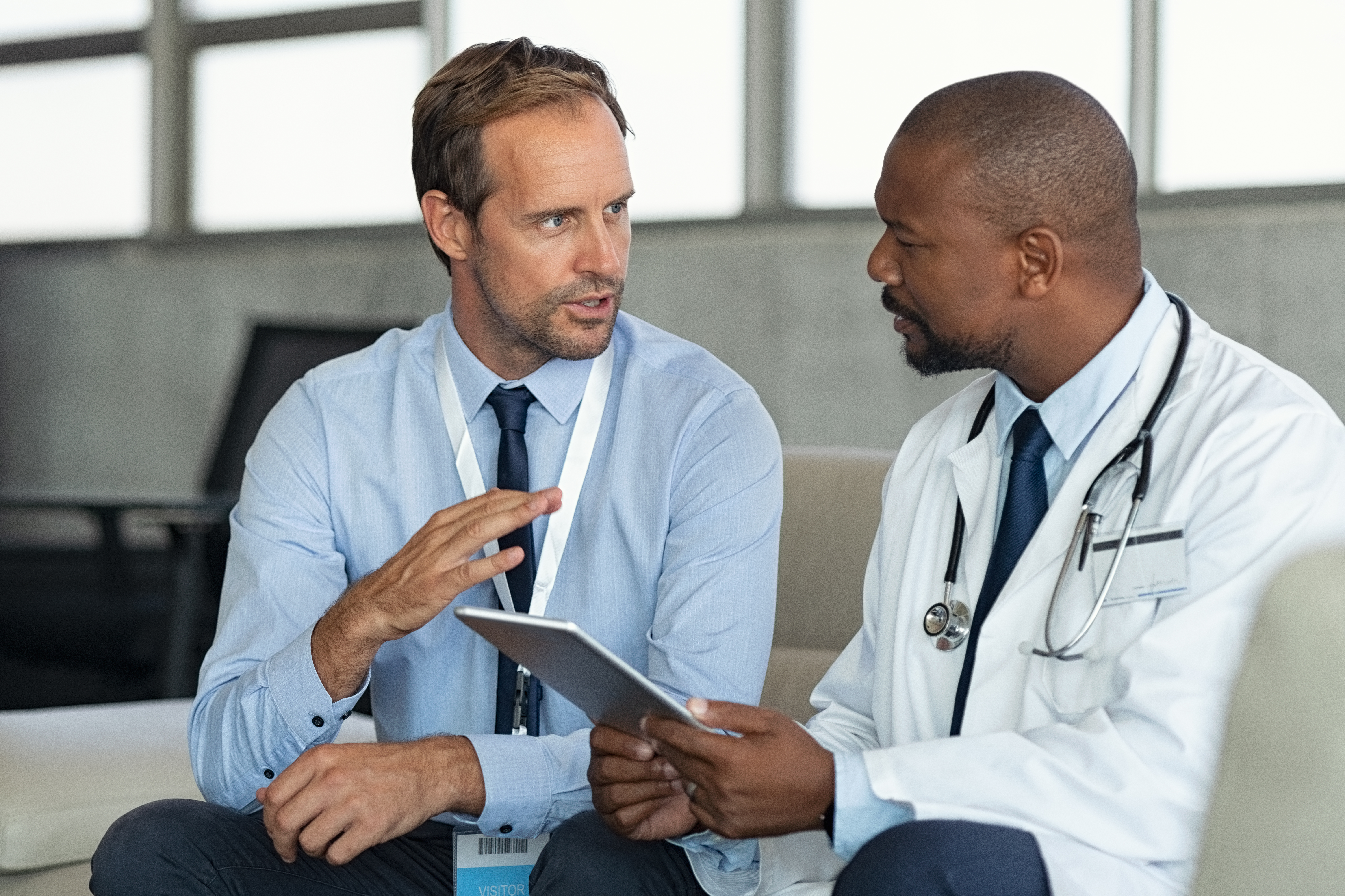 Pharmaceutical representative talking with doctor sitting on couch. African mature practitioner discussing results of the analysis with specialist while consulting diagnosis on digital tablet. Doctor in conversation with medical advisor in hospital room discussing patients 